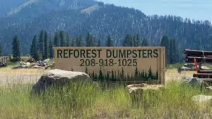 Reforest Dumpsters roadside sign with the company logo and phone number set against Idaho mountains and pine forest
